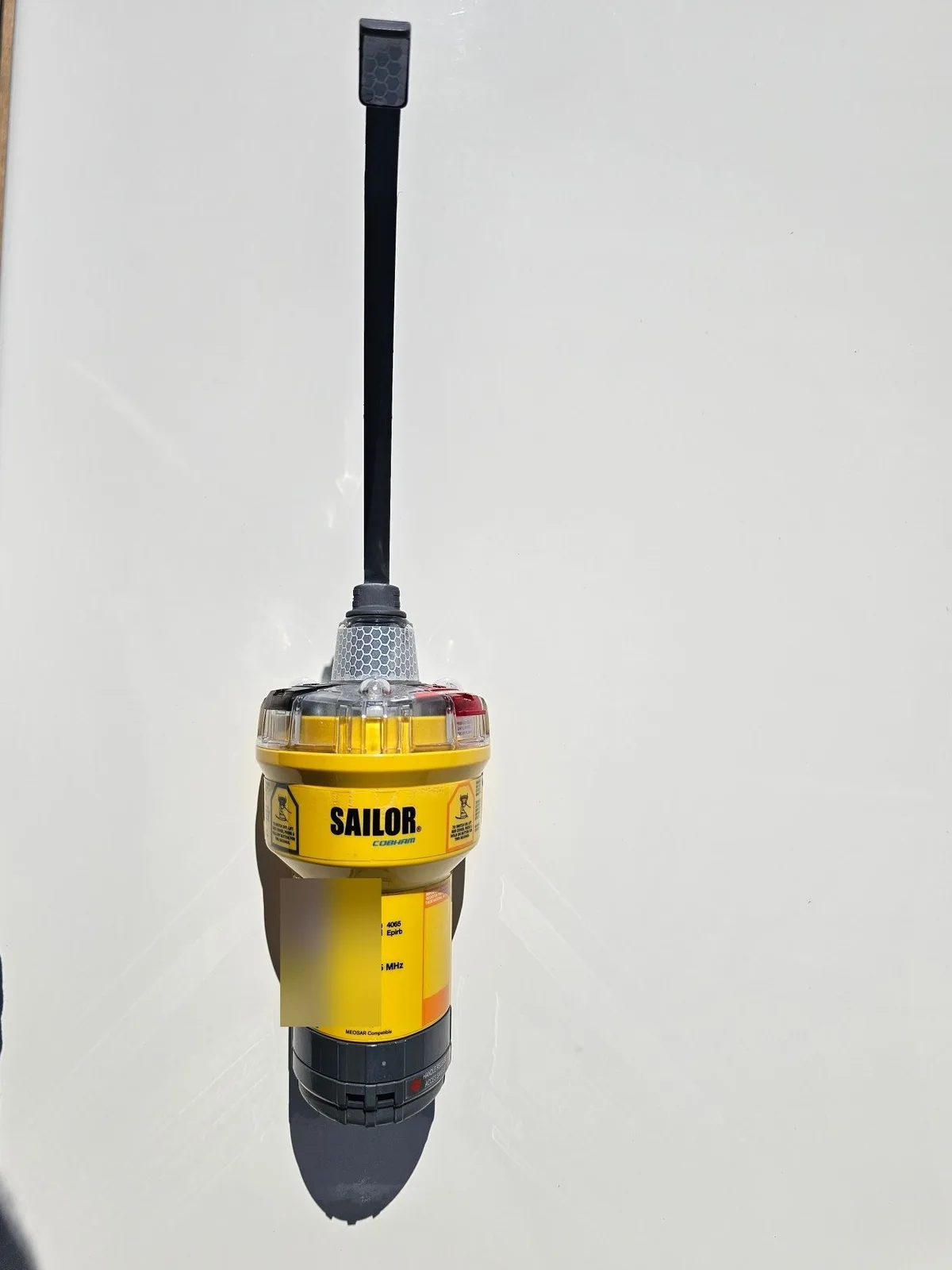 A yellow EPIRB with its aerial, laid on a white surface. A small label on the body is blurred.