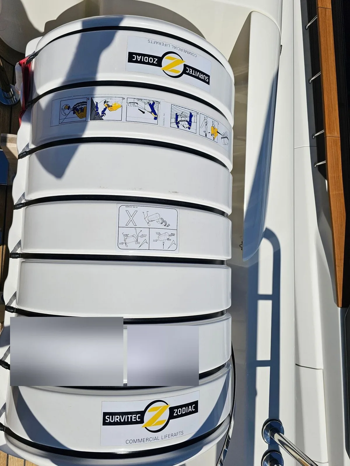 A white liferaft canister in its cradle on deck, seen from above, with picture labels on the casing. Two labels are blurred.