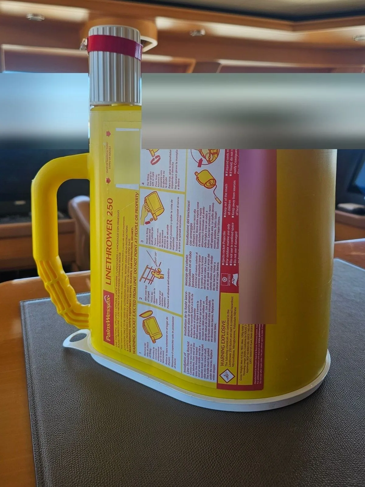A yellow linethrower standing on a table, with the maker's label and printed steps on the casing. The dates, lot number and the view through the windows are blurred.